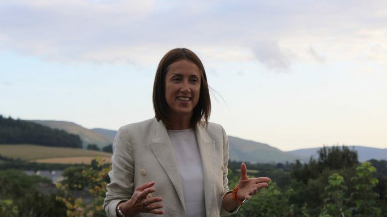 Jane Dodds in a white suit jacket and jeans in front of countryside